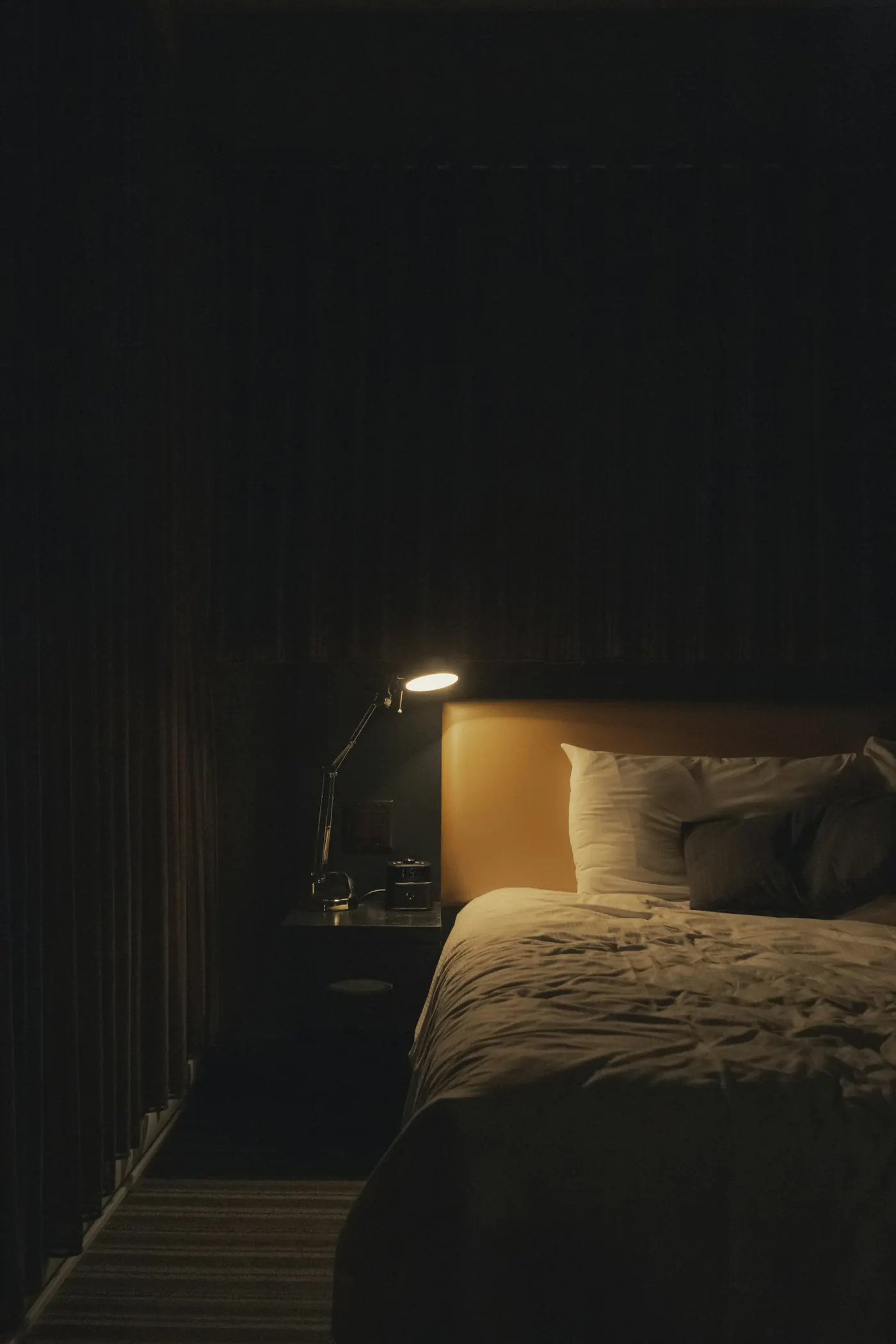 A dark bedroom at night, lit only by a small reading lamp beside the bed