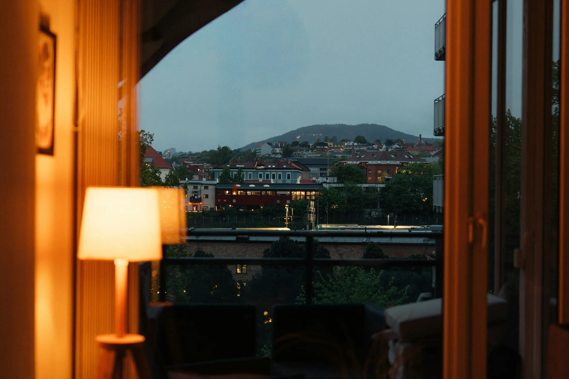 A lit table lamp by a window in the evening, with the city and a hill outside in blue light
